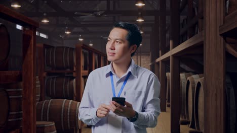 asian male professional worker standing with his smartphone in cellar of winery, he observes by looking around before he come to concentrating with his smartphone