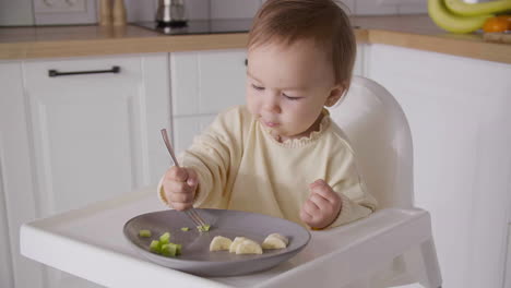 Cute-Baby-Girl-Sitting-In-High-Chair-In-The-Kitchen-And-Eating-Fruit-Slices-Using-Fork