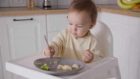 linda niña sentada en una silla alta en la cocina y comiendo rodajas de fruta usando un tenedor