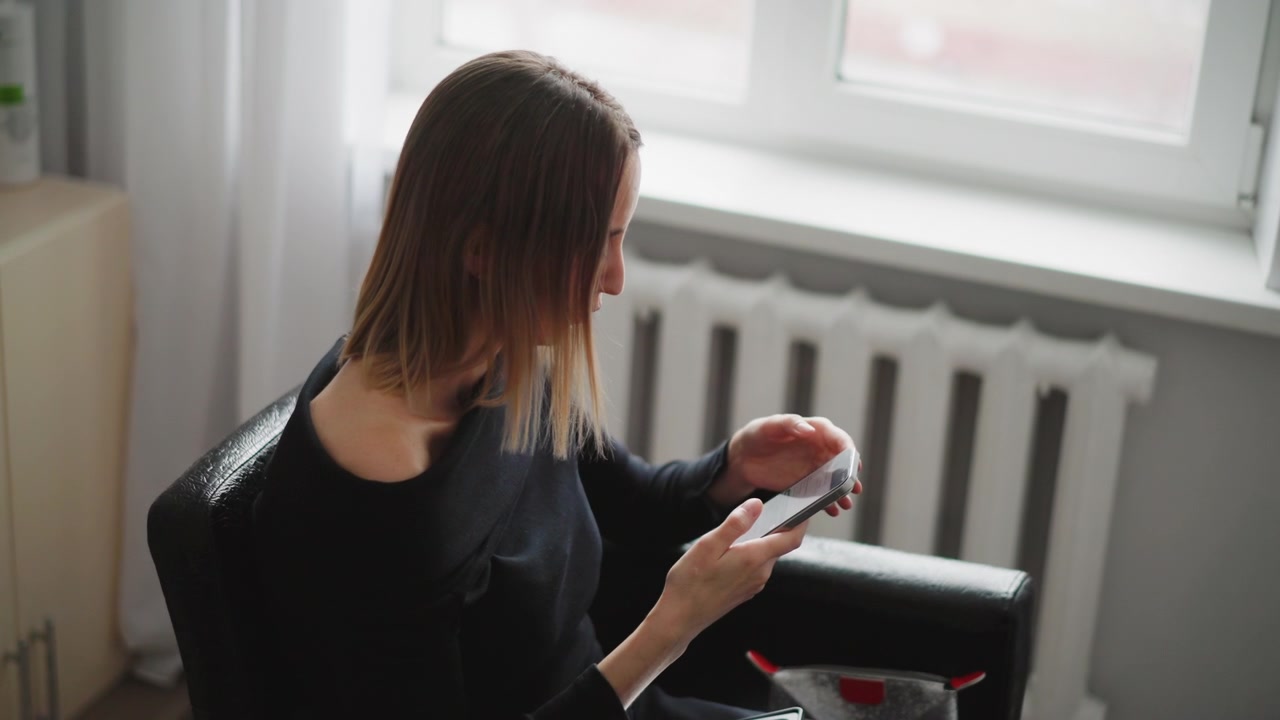 Nærbillede af kvinde, der bruger sin smartphone indendørs med blødt naturligt lys. Kvinden holder enheden i begge hænder og fokuserer på skærmen, mens hun sidder nær et vindue og en radiator. Scenen udstråler ro.