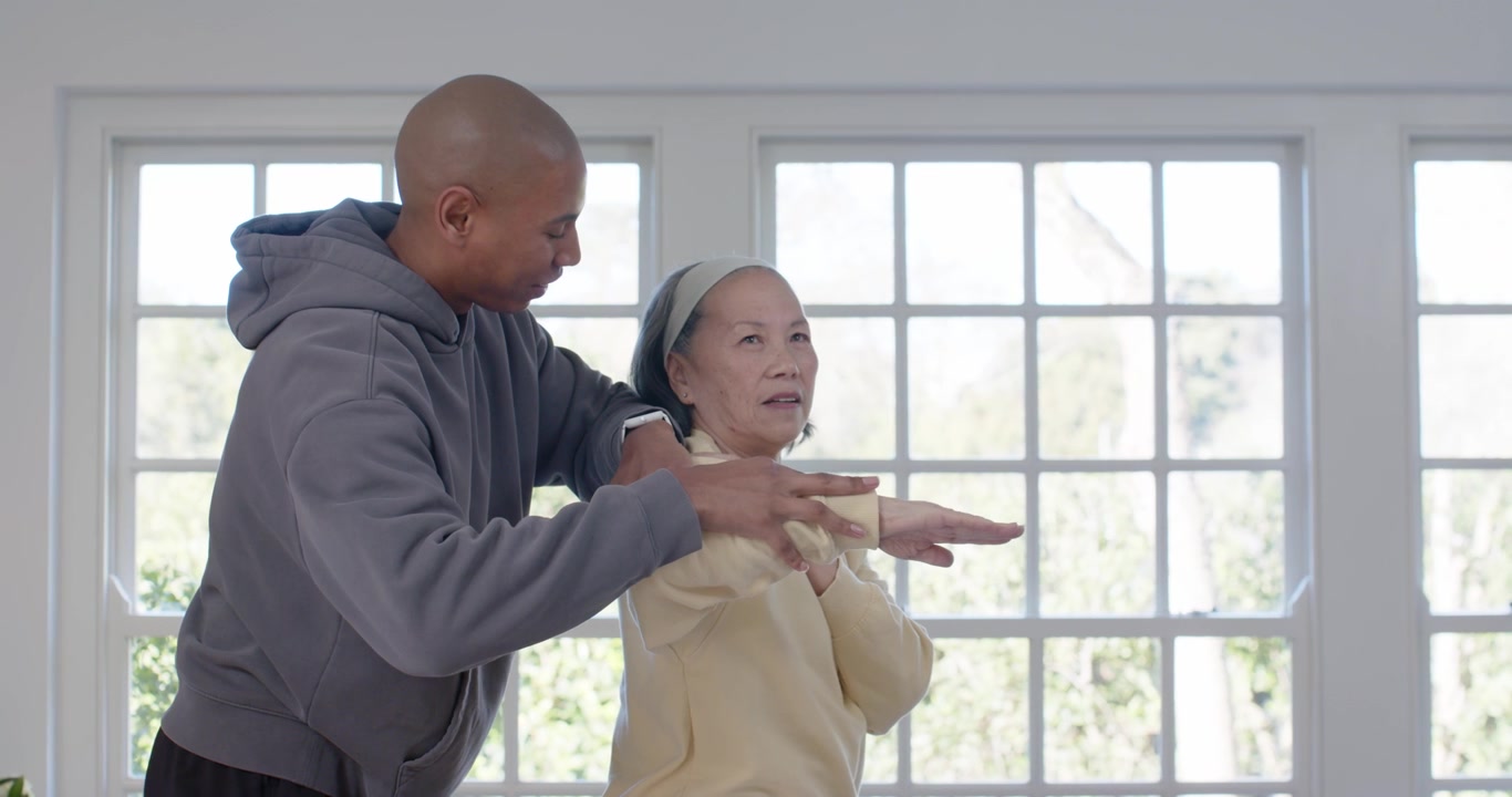 Adult man reaching under Senior Asian woman's arm, guiding shoulder stretch for mobility by window