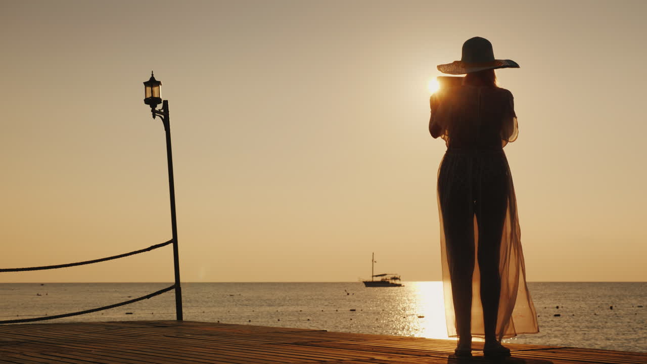 해변 옷을 입은 젊은 여성이 부두에 서서 아름다운 일출 사진을 찍고 있다