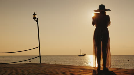 una joven vestida de playa está parada en el muelle y está tomando una foto de un hermoso amanecer