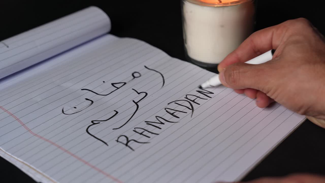 Lähikuva käsistä kirjoittamassa tekstiä ”Ramadan Kareem” arabiaksi ja englanniksi viivoitetulle muistilehtiöpaperille, tussikynä liikkeessä, lämmin kynttilänliekin hehku, tumma pöytä, jossa on tilaa tekstille, islamilaisen pyhän kuukauden tervehdys