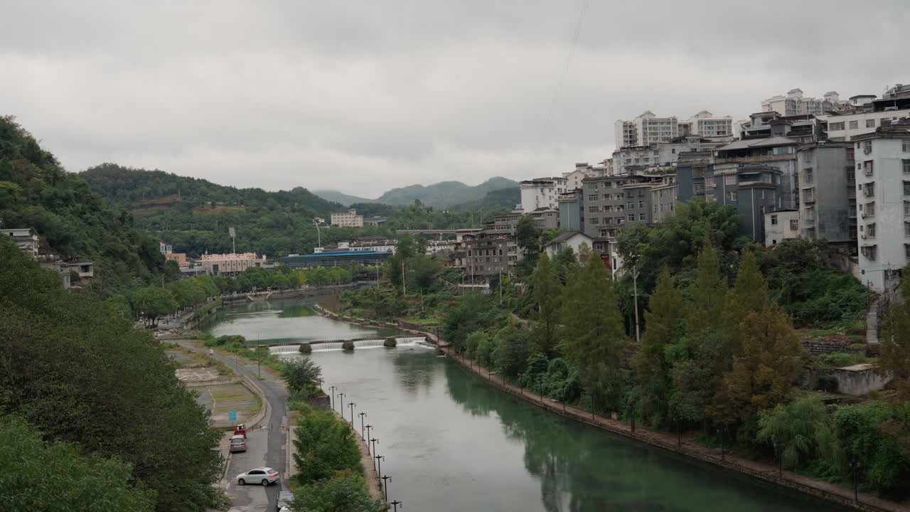 Valle fluviale nuvolosa con una città lungo il fiume, cielo grigio, condomini a più piani su un ripido pendio, riva del fiume alberata, strada stretta con traffico leggero, ponte distante e colline nebbiose. Atmosfera calma e riflessiva.
