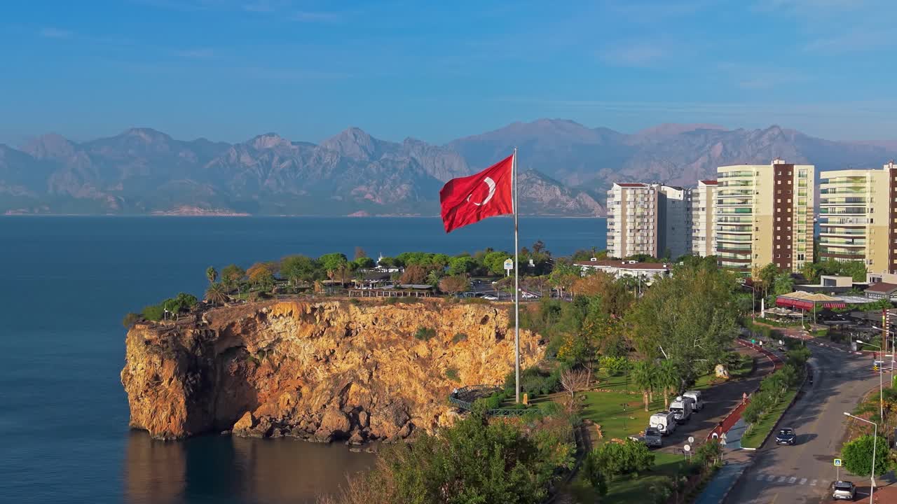 Antalya, Türkiye'deki Denizin Yüzü yakınlarında, kayalık bir sahilin, denizin ve yakınlardaki şehir binalarının üzerinde dalgalanan Türk bayrağının ağır çekim havadan çekimi; uzakta dağlar görünüyor.