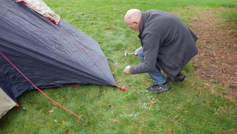 Ein-Mann,-Der-Mit-Einem-Hammer-Zeltheringe-In-Ein-Feld-Auf-Dem-Gras-Einschlägt,-Während-Er-Auf-Einem-Campingplatz-Campt