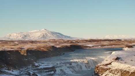 gullfoss waterfall in iceland cascades down the hvítá river, surrounded by a snow-covered landscape with a majestic mountain in the background under a clear blue sky.