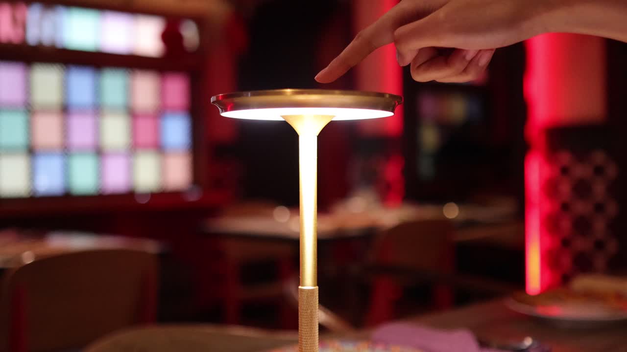 A hand activates a sleek, touch-sensitive table lamp in a dimly lit, colorful Bangkok café. Warm ambient lighting and shallow depth of field enhance the modern, cozy atmosphere