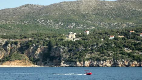 Vista-De-Colinas-Y-Acantilados-En-Croacia-Sobre-El-Agua-Del-Océano-Con-Un-Pequeño-Bote-Rojo-Navegando-Por