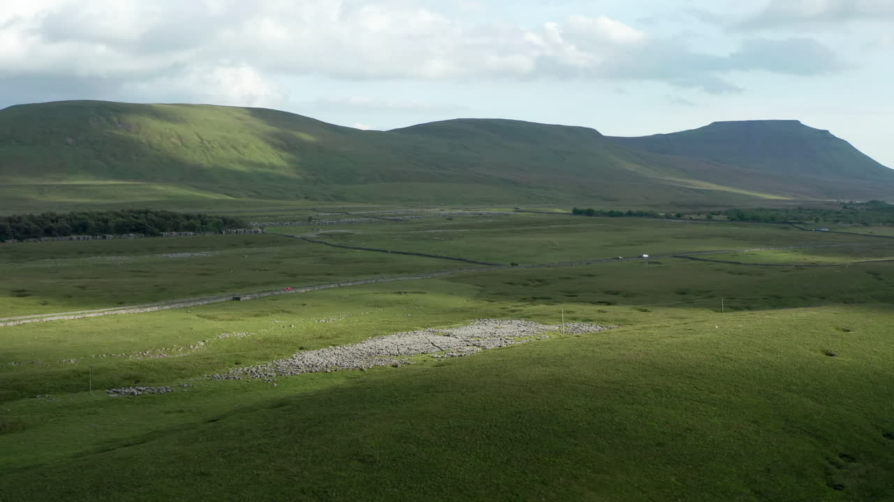 전경에 석회암 포장 도로가 있는 요크셔 데일스의 잉글버러 산 앞을 지나가는 자동차의 공중 돌리 샷