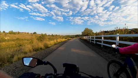 Radfahrer,-Die-Einen-Naturpfad-Entlang-Eines-Flusses-Hinunterfahren,-Wobei-Sich-Die-Morgensonne-Und-Der-Himmel-Vom-Wasser-Spiegeln---First-Person-Ansicht