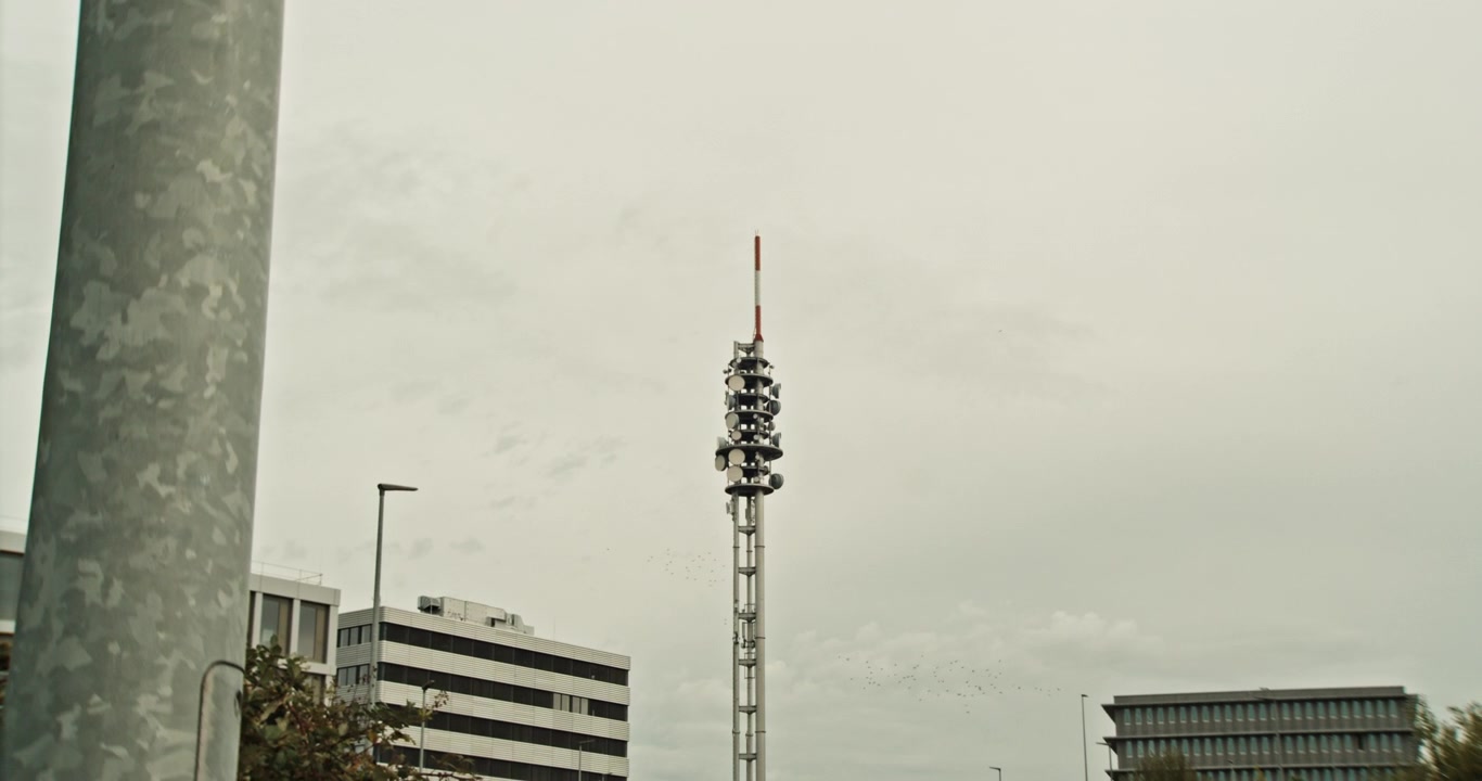 La telecamera si allontana da una vista medio-ampia di una torre di telecomunicazioni fino a un'inquadratura estremamente ampia, con l'antenna che diventa piccola contro il cielo e l'ambiente circostante. Infrastruttura di comunicazioni, Germania