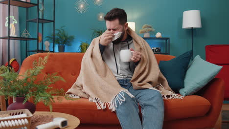 sick ill man wrapped in plaid sit alone shivering from cold on sofa drinking hot tea on home sofa