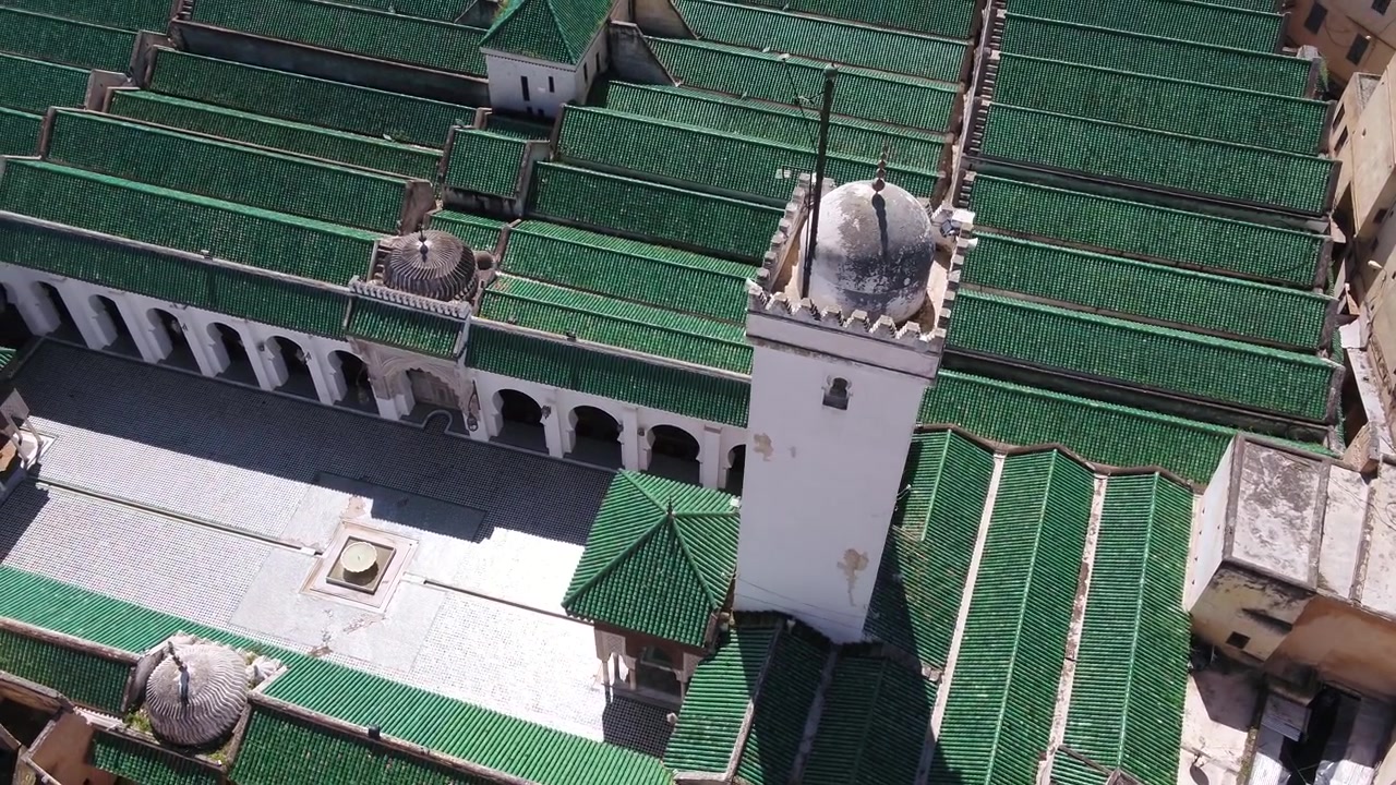 Ripresa aerea con drone ad alta angolazione che cattura lo storico minareto bianco, il bordo del cortile e i tetti paralleli in tegole verdi della Moschea Al Quaraouiyine a Fez, Marocco