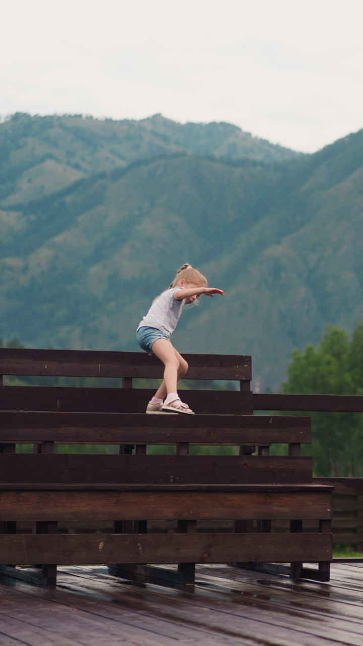 Little girl climbs on wet wooden benches while young brother walks along large dancing ground after drizzling rain against old mountains slow motion