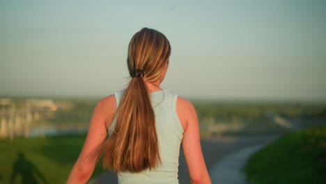 back view of lady roller skating on path with long hair tied back, under warm sunlight, shadow cast on ground, with blurred cityscape and greenery in background