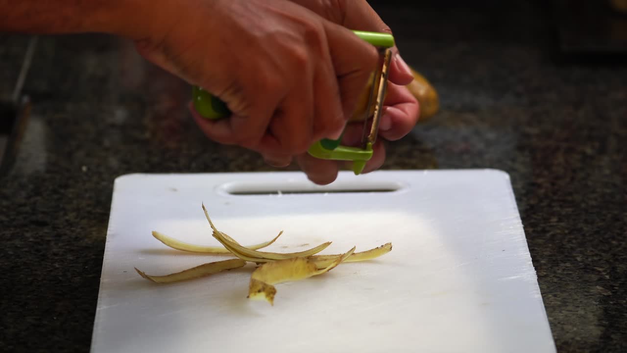 Middle aged brown skin man peeling potatoes with peeler on a white chopping board at home