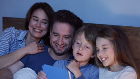 Vista-De-Cerca-De-Los-Padres-Caucásicos-Sentados-En-La-Cama-Con-Su-Pequeño-Y-Lindo-Hijo-E-Hija,-Viendo-Algo-En-La-Tableta-Y-Hablando-Por-La-Noche