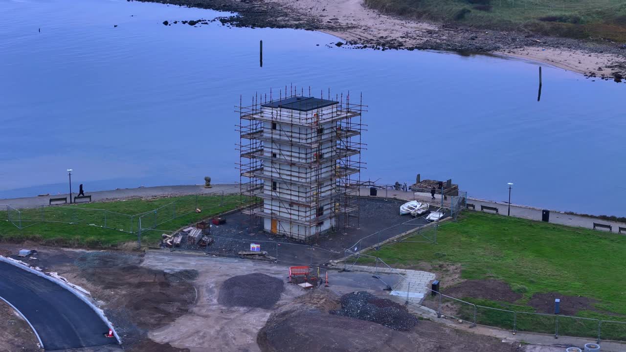 Aerial view captures coastal construction site at sunset, showing scaffolding around an old lighthouse structure near the shoreline and walking paths in a tranquil waterfront zone