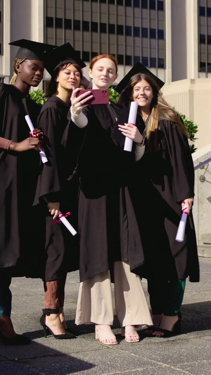 Video verticale: Inclinando un telefono rosa, una ragazza dai capelli rossi si scatta un selfie con i colleghi in toga in piazza con i diplomi.
