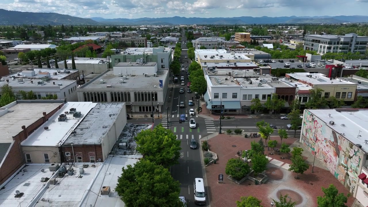 Medfords hovedgade strækker sig forbi et farverigt vægmaleri og en skyggefuld plads, med trafik ved fodgængerfeltet og fjerne skovklædte bjerge, der indrammer en overskyet forårshorisont. Oregon, USA. Baglæns droneoptagelse.