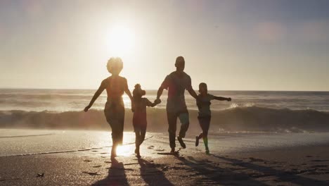 una familia afroamericana sonriente corriendo y tomándose de las manos en una playa soleada