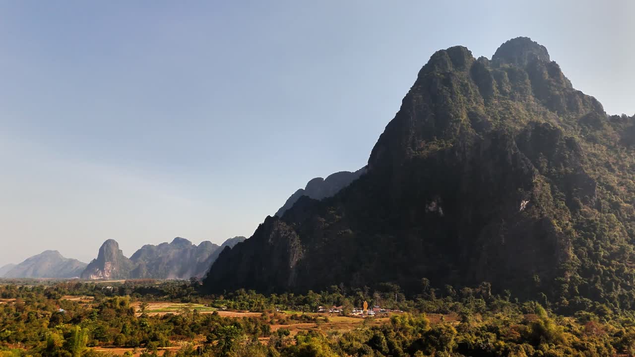 Low drone pan in Vang Vieng, Laos showing towering karst cliffs, farmland, and scattered rural homes beneath rugged limestone peaks under soft morning light in dry season