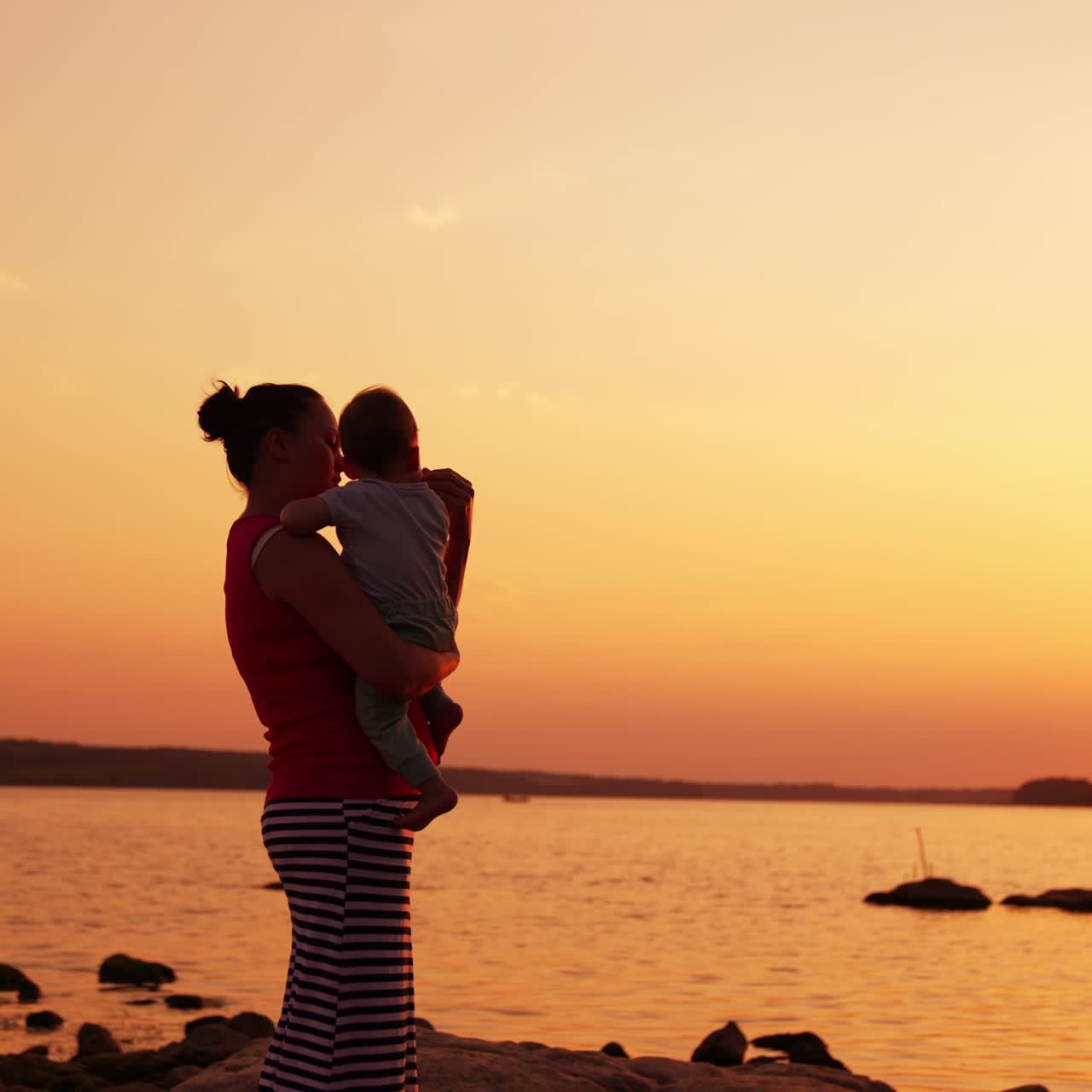 Caring and loving mother with her little baby in hands standing near the river. Family enjoying time at the nature at sunset