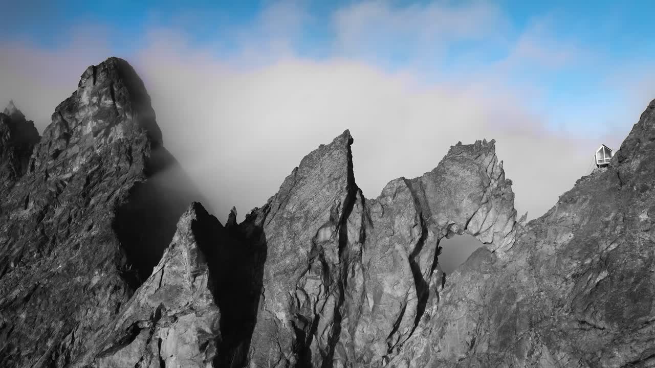 Aerial drone footage flying over and above Western alps dark gray mountains in Valle D'Asota during a sunny day with a small mountain climbers cabin on the steep edge and blue sky in the background.
