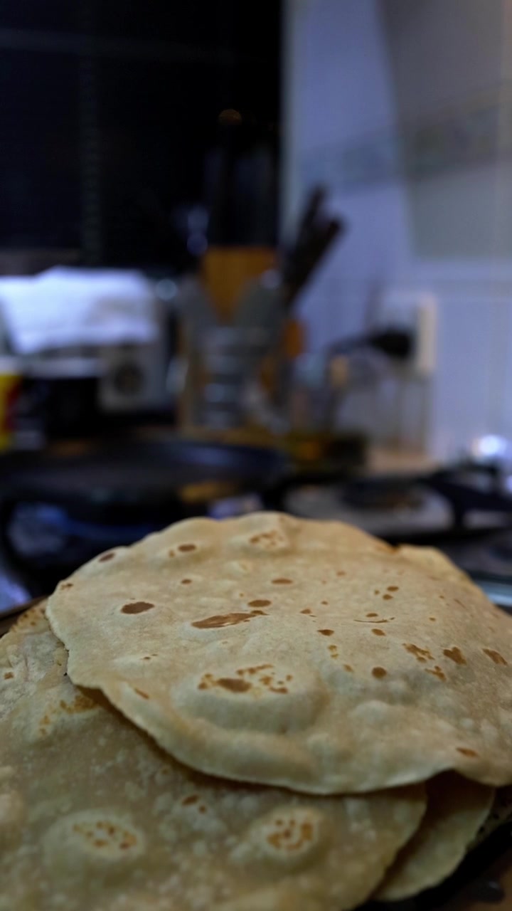 Close-up of golden-brown chapati being cooked on a dark skillet in a kitchen. A person's hand holds a metal spatula, flipping the traditional Indian flatbread with visible char marks. Warm, focused li