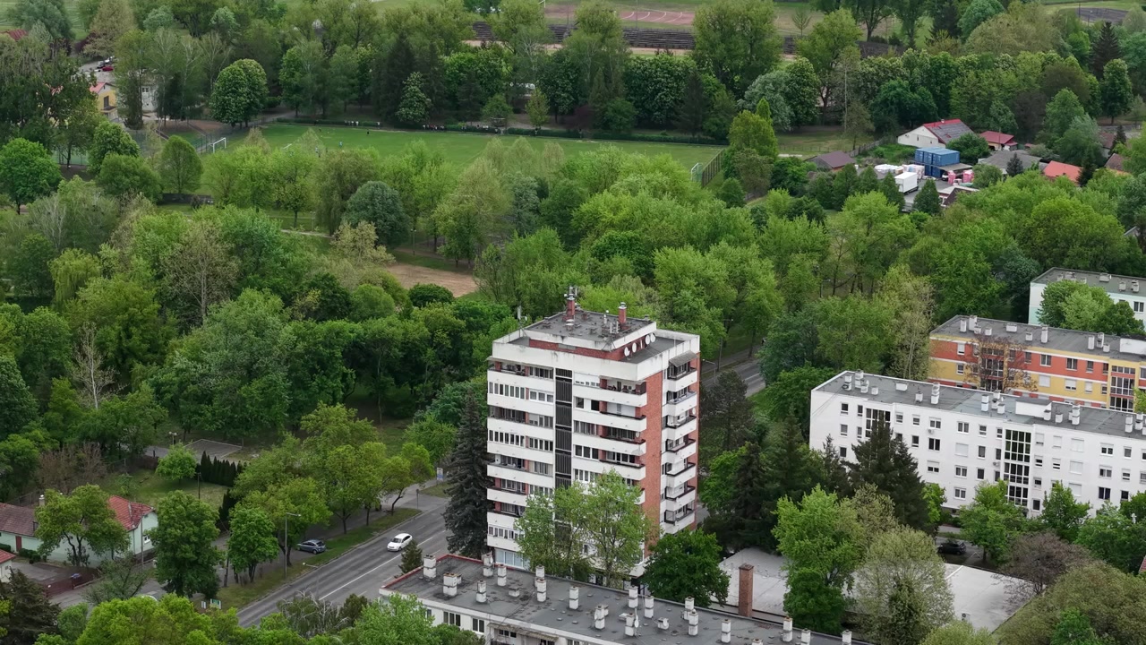 Pohled z ptačí perspektivy ukazuje moderní obytný bytový komplex obklopený svěžími jarními stromy poblíž klidné městské ulice s provozem