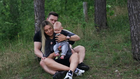 blonde woman sits leaning on her husband and holding a baby. beautiful caucasian family having good time in nature.