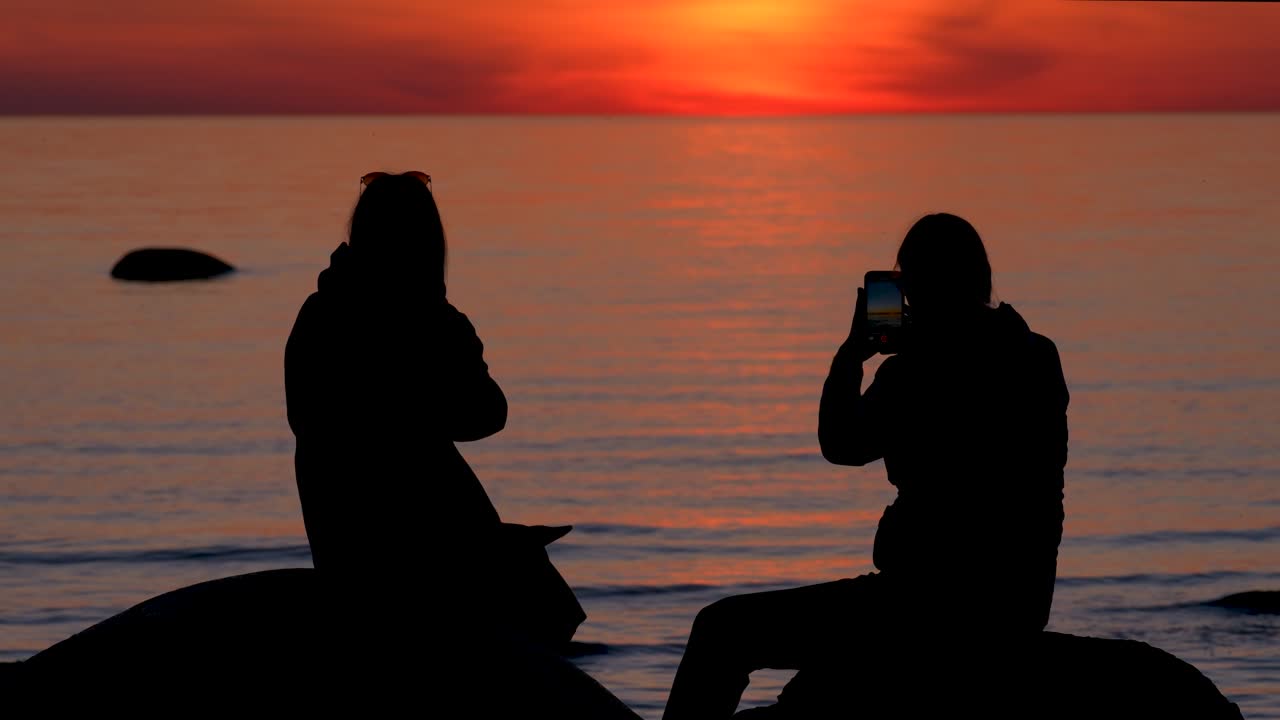 Young women silhouettes sitting by water during sunset.