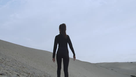 Mujer-Misteriosa-Vestida-De-Negro-Por-La-Espalda-Mirando-El-Desierto-Mientras-El-Viento-Sopla-Fuerte-En-Cámara-Lenta