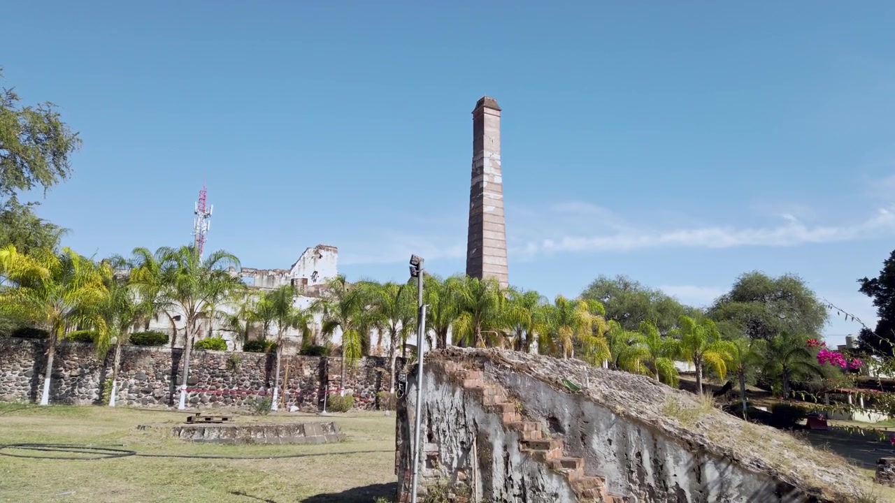 Vista dal basso dello storico camino in mattoni dell'Hacienda Coahuixtla, circondato da palme e terrazze in pietra sotto un cielo azzurro limpido a Morelos, Messico