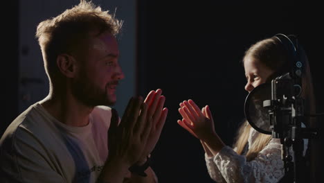 a young girl sings into a microphone in a recording studio, while a man claps and encourages her.