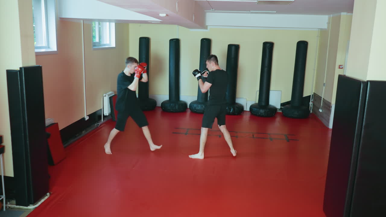 무술 체육관 안에서 스파링을 연습하는 운동선수들, 한 명은 발차기를 하고 상대는 빨간색 장갑을 끼고 전투 태세를 갖춘 채 빨간색 매트 바닥에서 훈련 세션 동안 키 큰 샌드백으로 둘러싸여 있어요