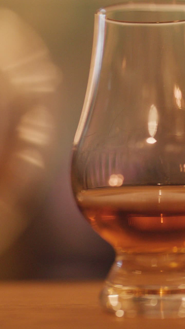Una mano fa roteare delicatamente un bicchiere di whisky, creando movimento nel liquido ambrato. Un'illuminazione calda e soffusa, simile a quella delle candele, e una messa a fuoco ridotta evocano un'atmosfera accogliente e intima.