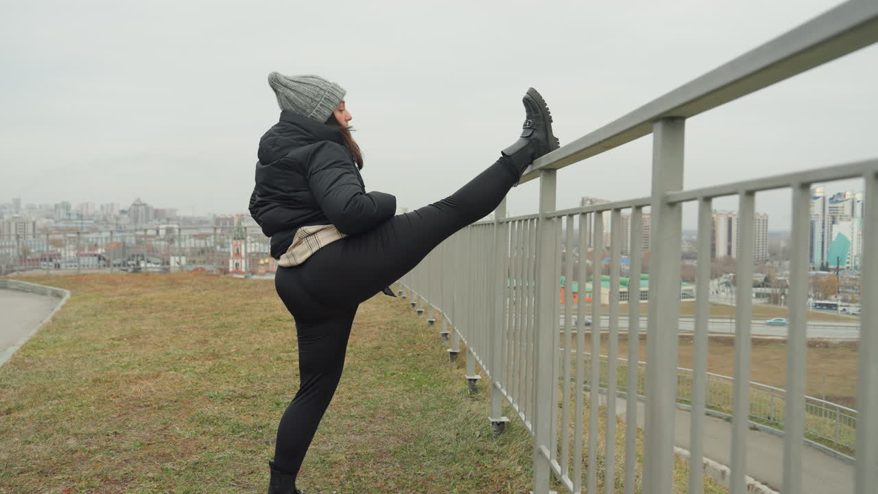 White woman stretching leg on railing, winter coat and beanie, overcast city skyline, slow controlled dance warmup and flexibility exercise, graceful kick and balance, urban rooftop overlook, focused