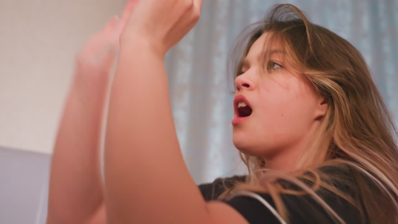 Side view of young brunette girl with flowing hair and calm expression as she playfully reacts with surprise during soft pillow hit in cozy bedroom