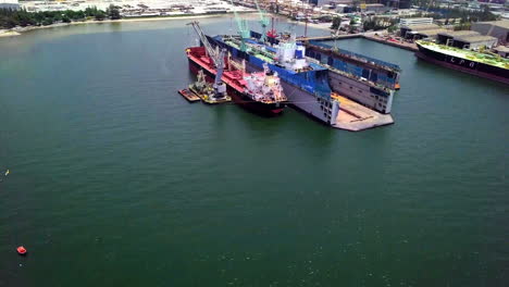 Aerial-view-of-logistics-concept-a-cargo-ship-being-retrofit,-repair-and-services-on-a-floating-dry-dock-off-the-coast-at-Laem-Chabang-dockyard-in-Chonburi-Province,-Thailand