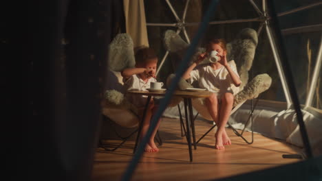 children with delicious drinks under glamping dome. little boy stirs beverage and girl sips from cup sitting at table in tent. evening tea at camp