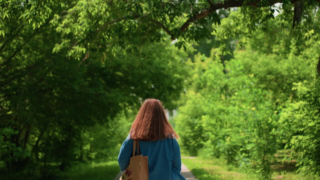 Back view of excited girl walking playfully along paved path in sunny park, dancing joyfully while holding embroidered fabric, wearing casual outfit and sneakers, surrounded by gentle sunlight