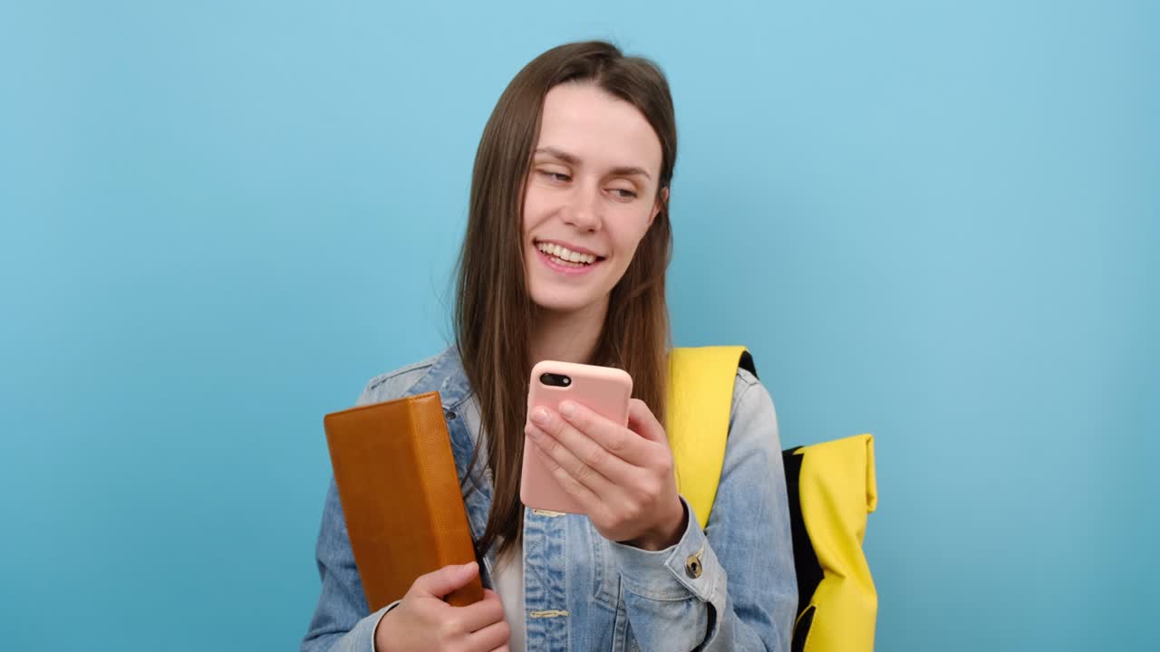 retrato de una chica adolescente con libros en la mano, usa un teléfono móvil, usa una chaqueta de denim y una mochila amarilla, aislado en la pared de fondo de color azul en el estudio. educación en el concepto de universidad