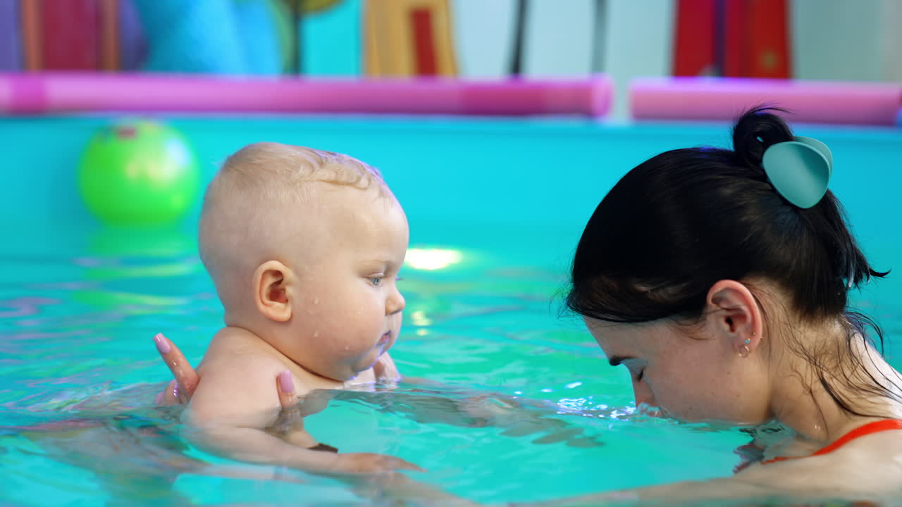 Adorable chubby blond baby in the swimming pool. Coach holding a child teaches the kid to hold air.