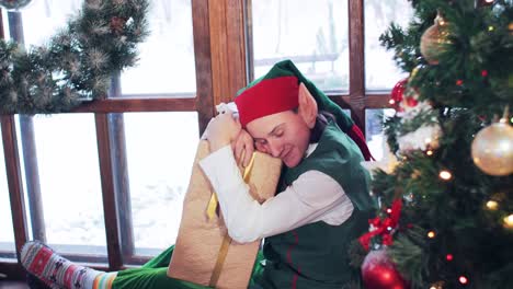 elf yawns and sits on a windowsill near a christmas tree with decorations hugging a box with gifts