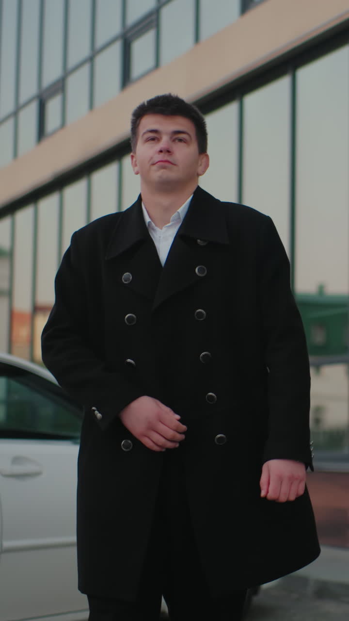 hombre con abrigo negro ajustando abrigo, poniendo la mano en el bolsillo, caminando majestuosamente a través del entorno empresarial urbano, caminata profesional con edificios de oficinas y coches estacionados en el fondo