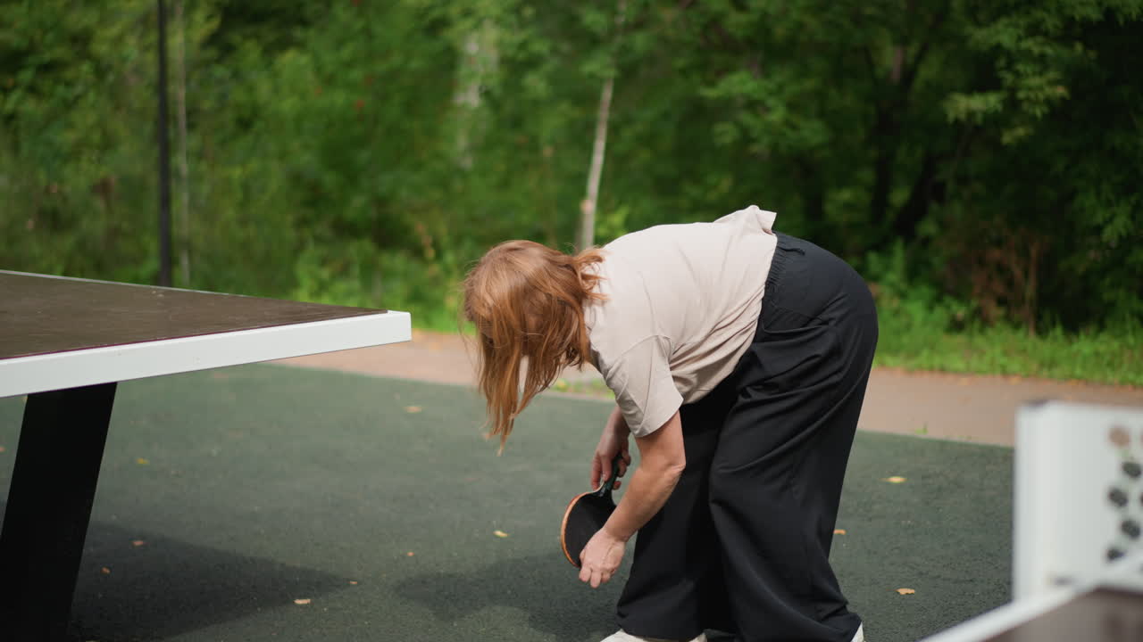 Retrieving Dropped Ball During Outdoor Match Caucasian Woman Bending To Pick Ball, Paddle In Hand, Quick Movement Across Park Table, Energetic Playful Vibe And Candid Action Capture