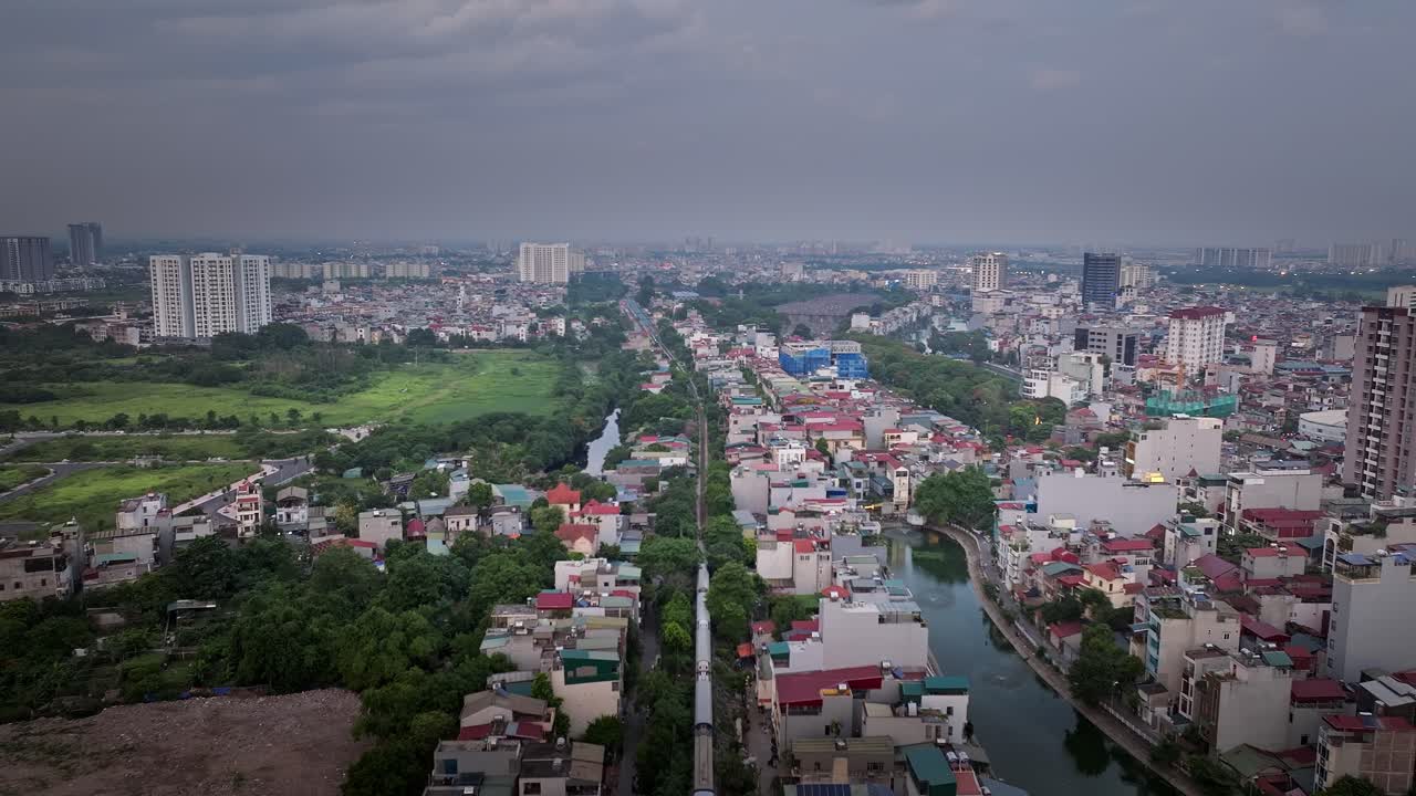 Aerial overview tracking follows train in Hanoi with tracks cutting through the city, capturing the mix of urban and green spaces, lively cityscape under cloudy sky
